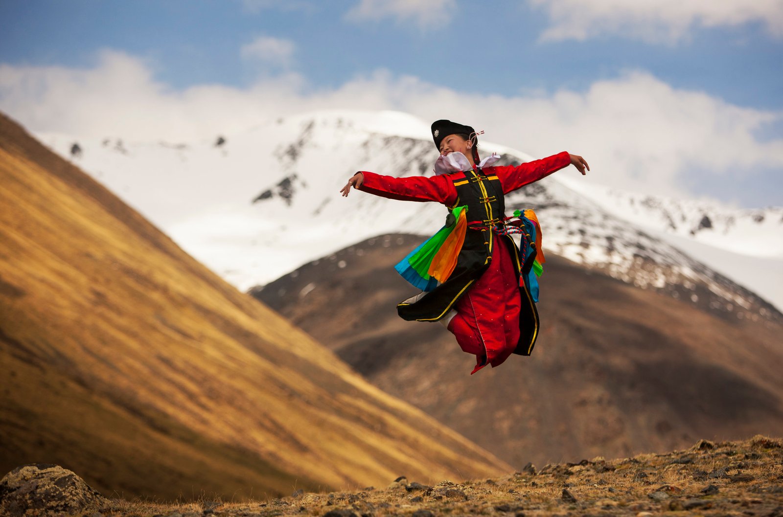 Dancer in a red deel leaping above a golden hillside below snow covered peaks