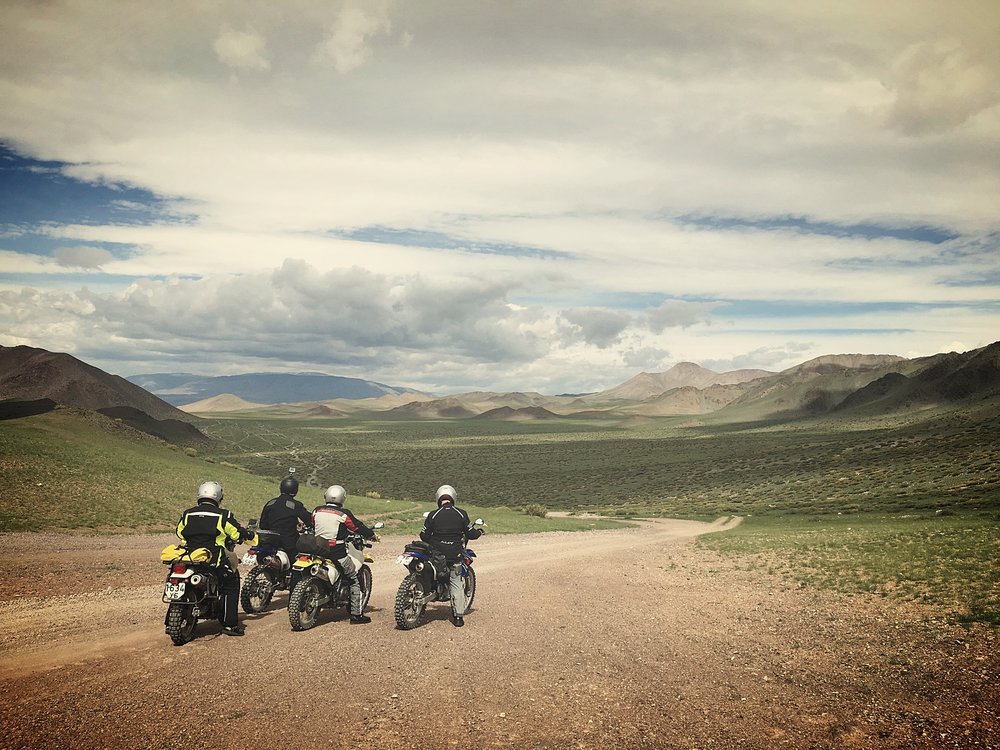 Three motorbikes pulled up on a dirt road above a wide green valley